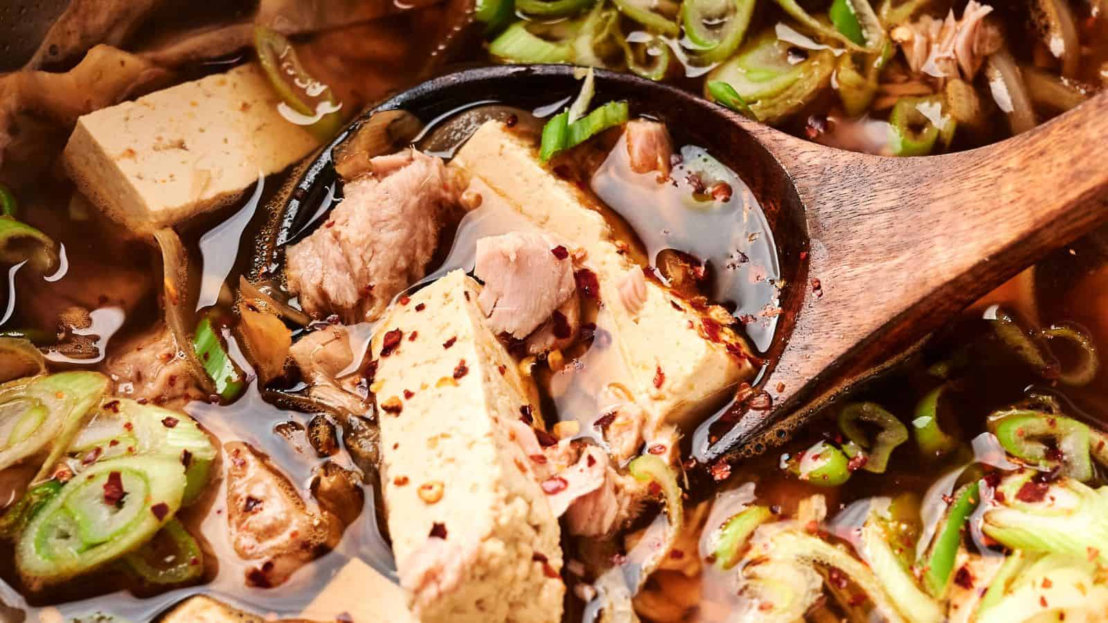 Close-up of a wooden spoon in a vibrant kimchi stew with tofu and pork, garnished with sliced green onions and red pepper flakes.