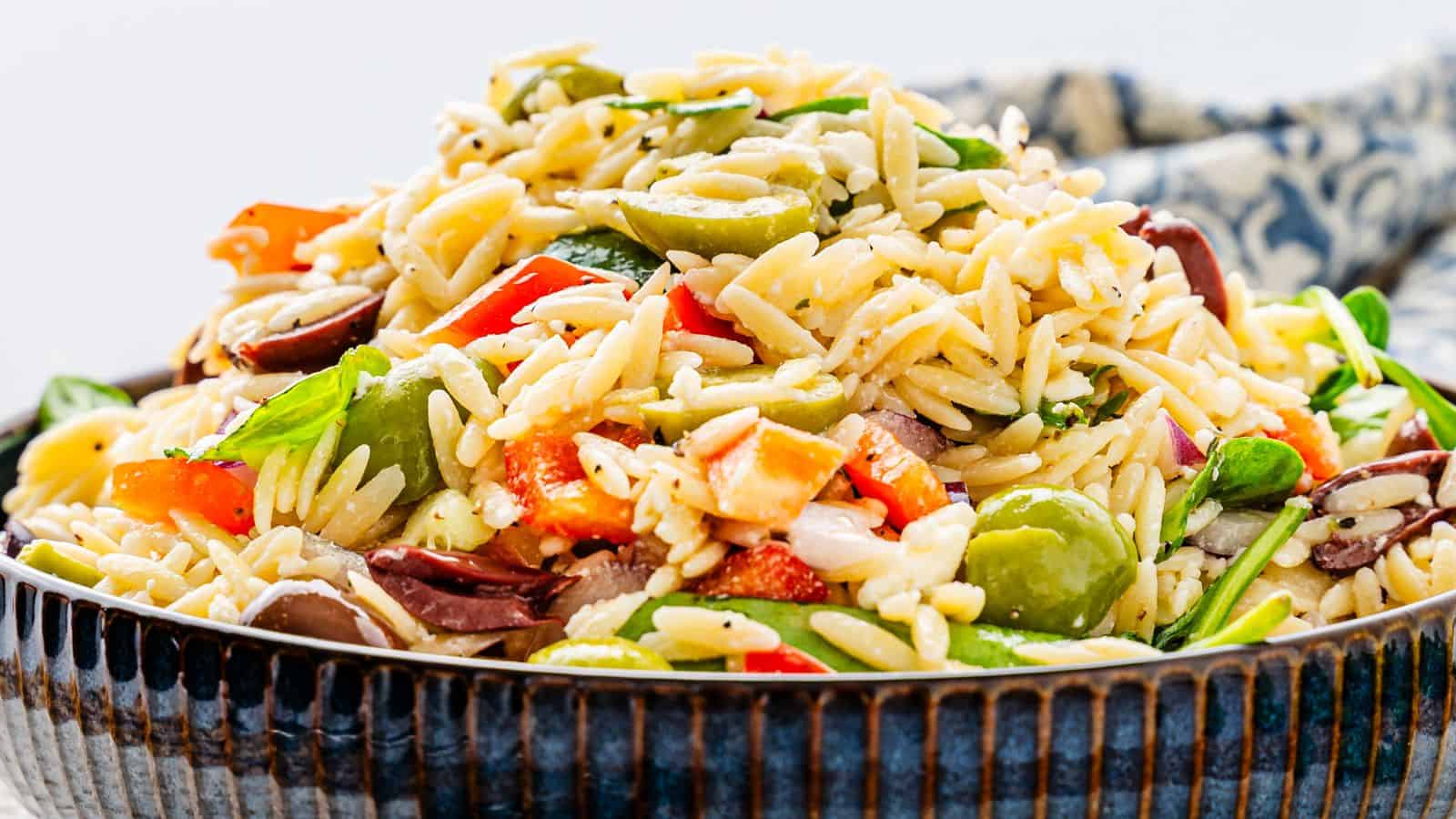 A bowl of orzo salad with mixed vegetables, including olives, tomatoes, and spinach, garnished with herbs.