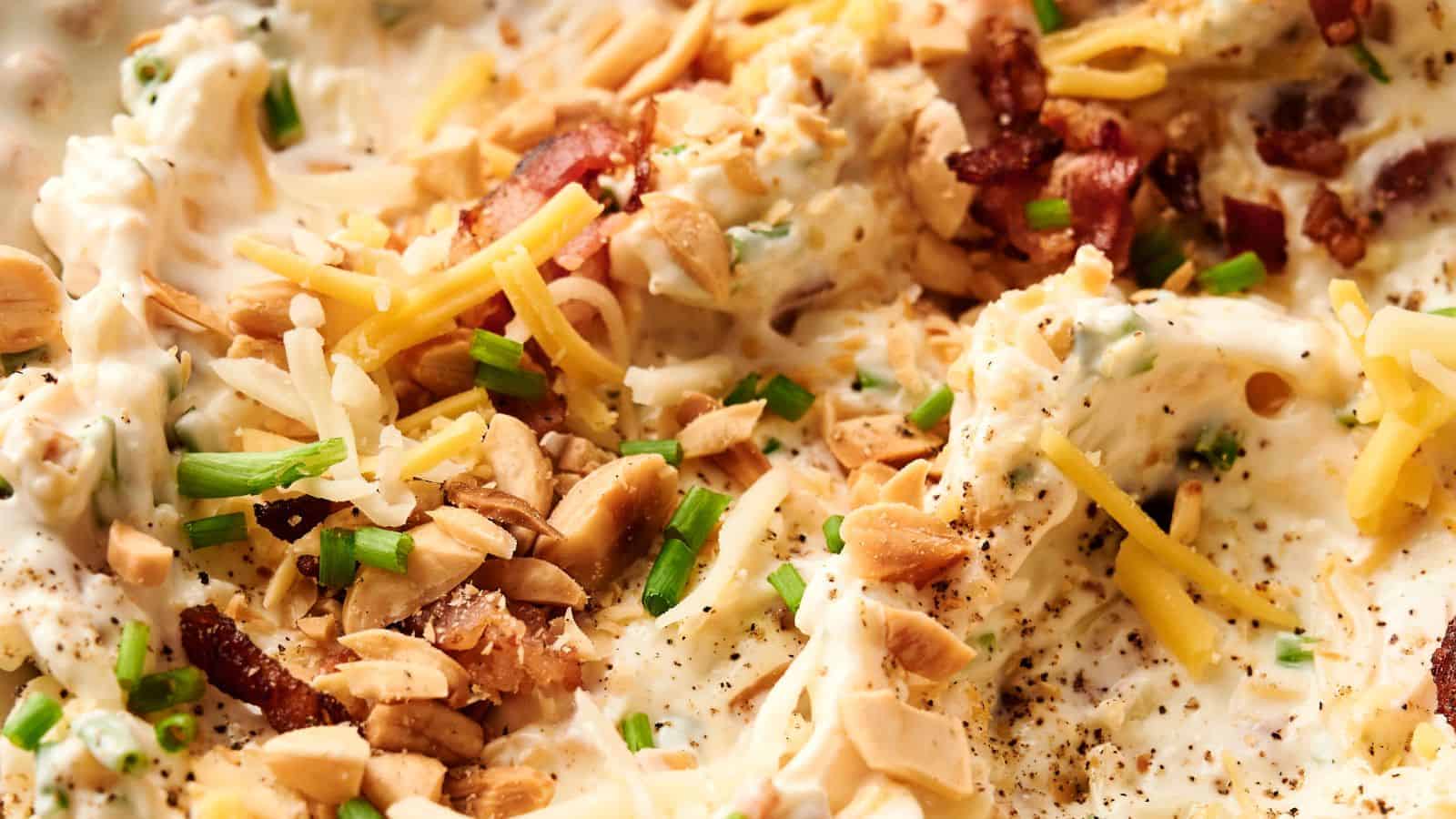 Close-up of a creamy dish topped with shredded cheese, chopped nuts, bacon bits, and sliced green onions.