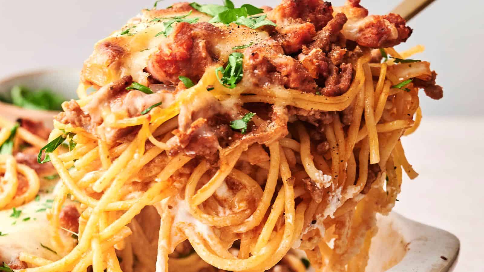 Close-up of a serving of cheesy baked spaghetti with meat sauce, topped with fresh parsley, being lifted from a dish.