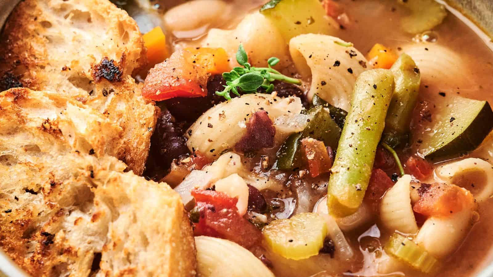 A bowl of hearty minestrone soup packed with green beans, pasta, zucchini, tomatoes, and herbs, served with crusty bread slices.