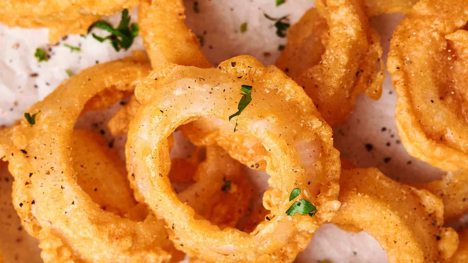 A close-up of crispy, golden-brown onion rings sprinkled with chopped parsley and black pepper on a white parchment-lined surface captures perfection in every bite.