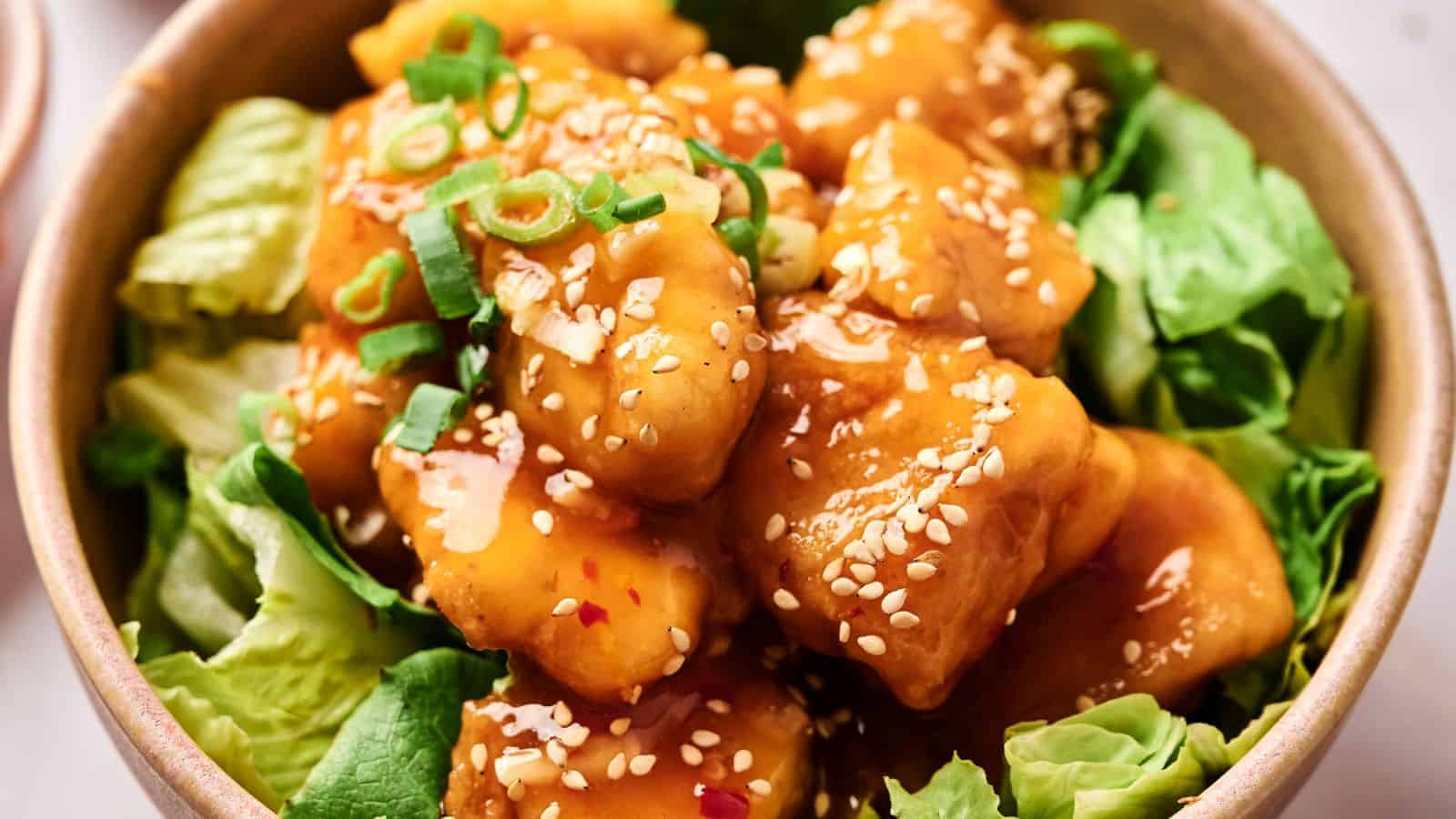 A bowl of crispy tofu coated in sesame sauce, garnished with green onions and sesame seeds, served on a bed of lettuce.