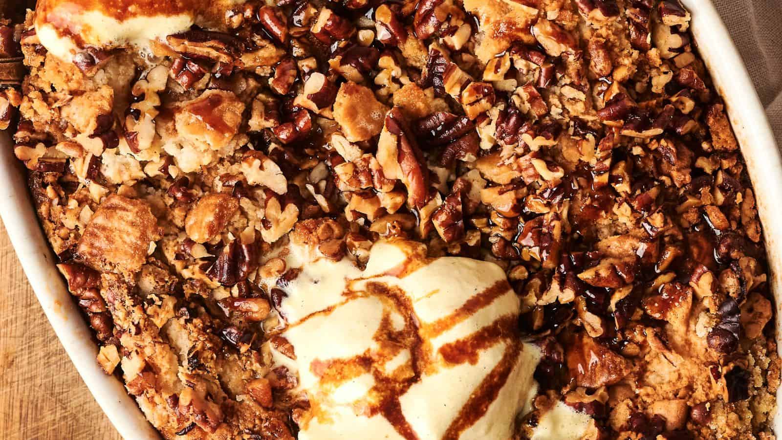 Close-up of a Pumpkin Dump Cake, a baked dessert topped with chopped pecans and drizzled with caramel sauce, served in a white dish on a wooden surface.