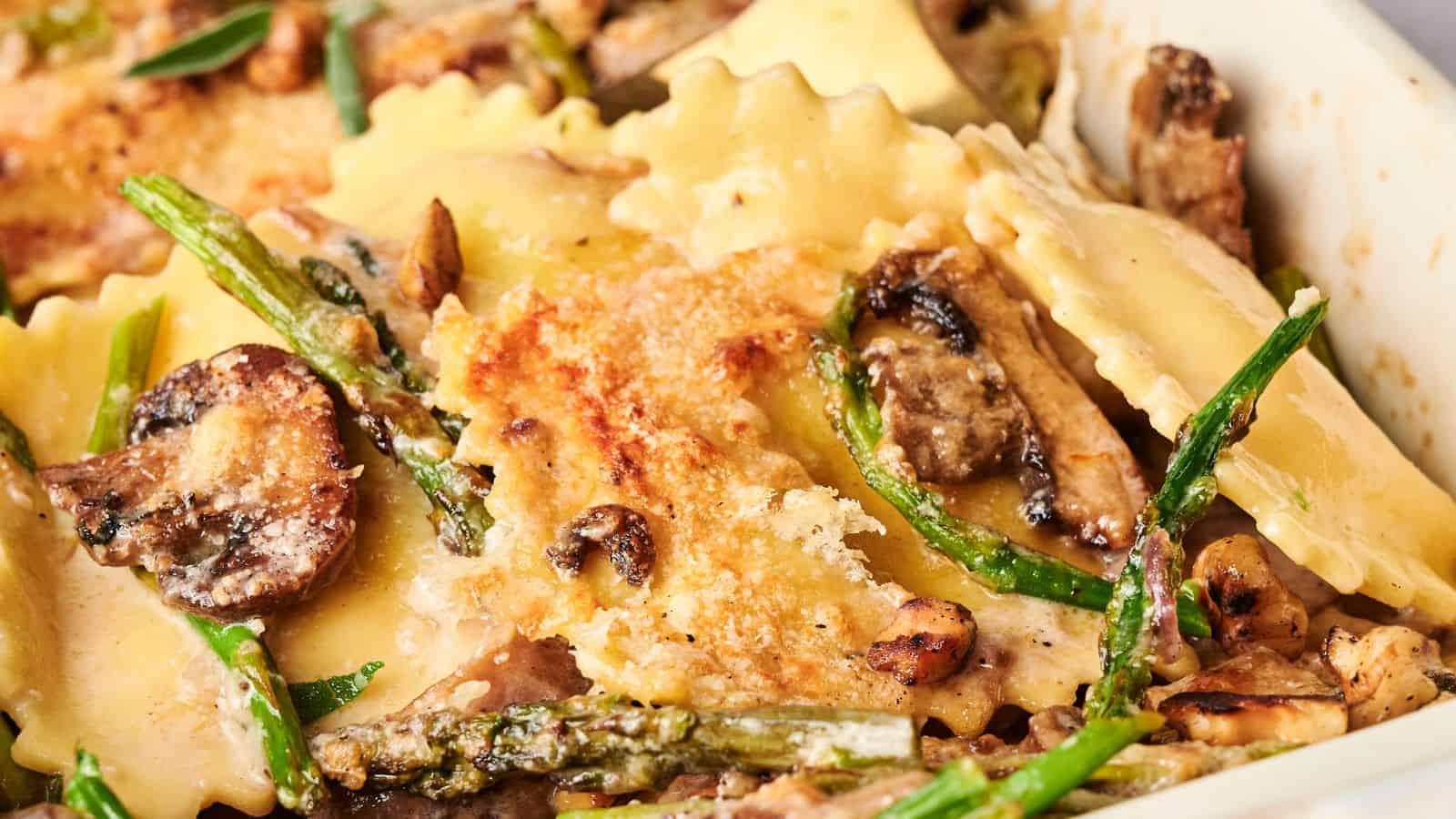 Close-up of a baked dish with ravioli, mushrooms, asparagus, and melted cheese on top.