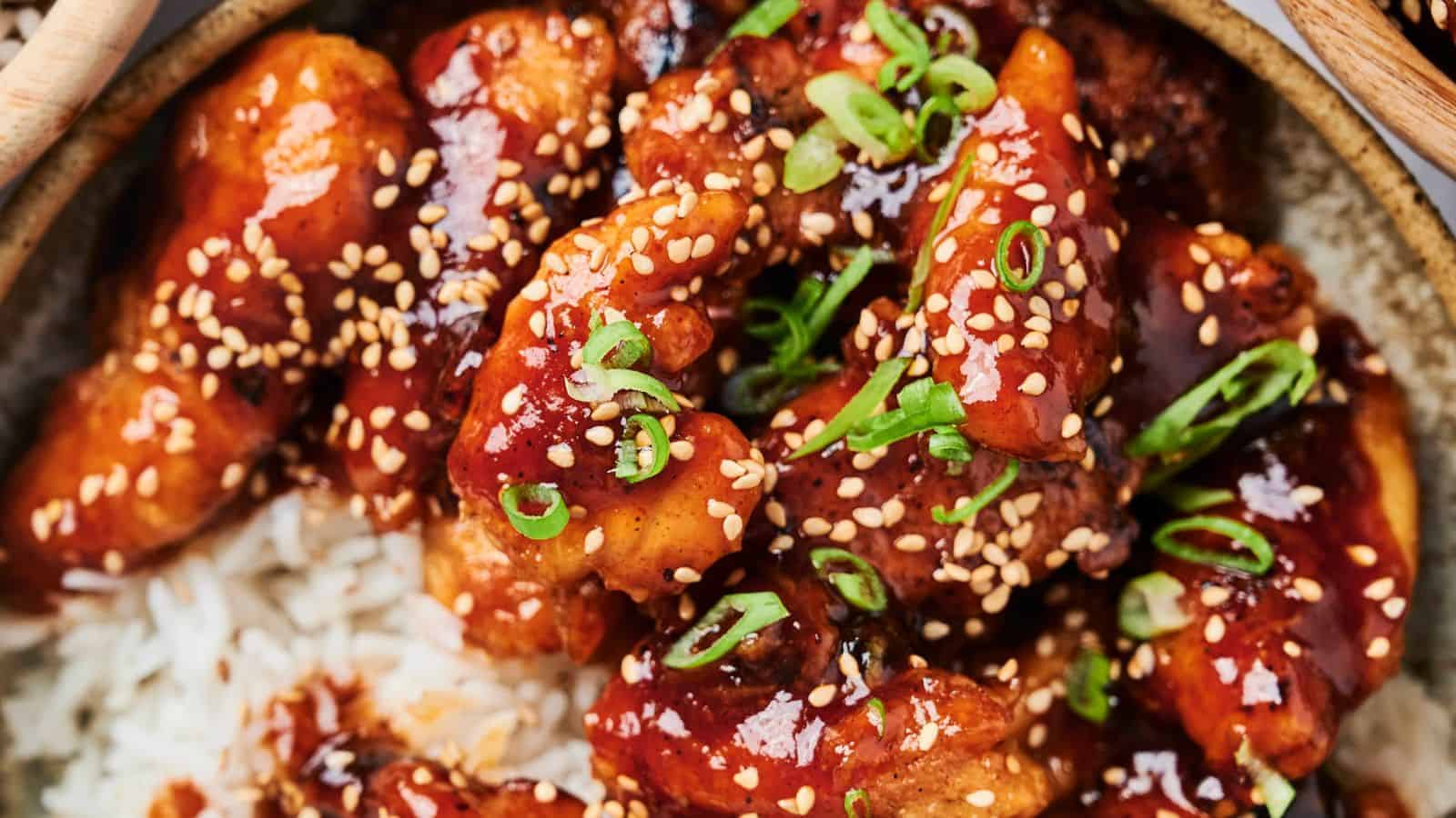 A close-up of glazed chicken pieces with sesame seeds and chopped scallions on a bed of rice.