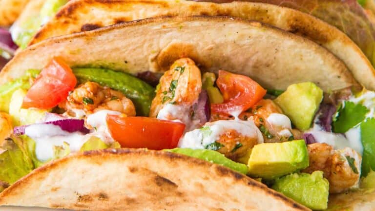 Close-up of a shrimp taco with avocado, tomato, red onion, and a drizzle of sauce in a toasted tortilla.