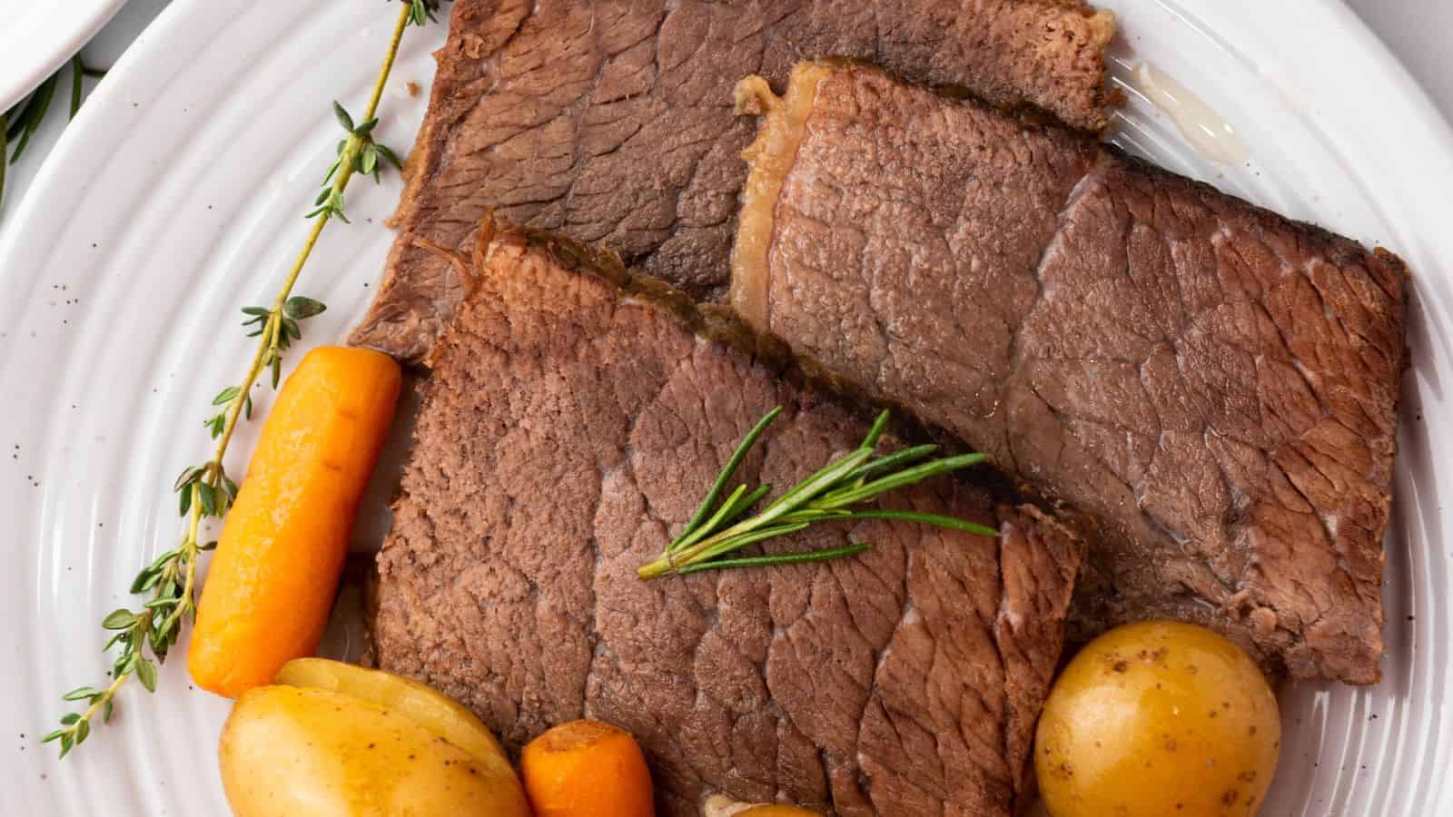 Slices of cooked beef with sprigs of herbs, served with small potatoes and carrots on a white plate.