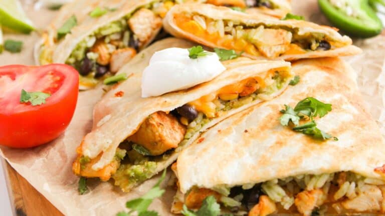 Quesadillas filled with chicken, beans, rice, and vegetables, topped with sour cream and garnished with cilantro. Sliced tomato and jalapeño pepper accompany the dish on parchment paper.