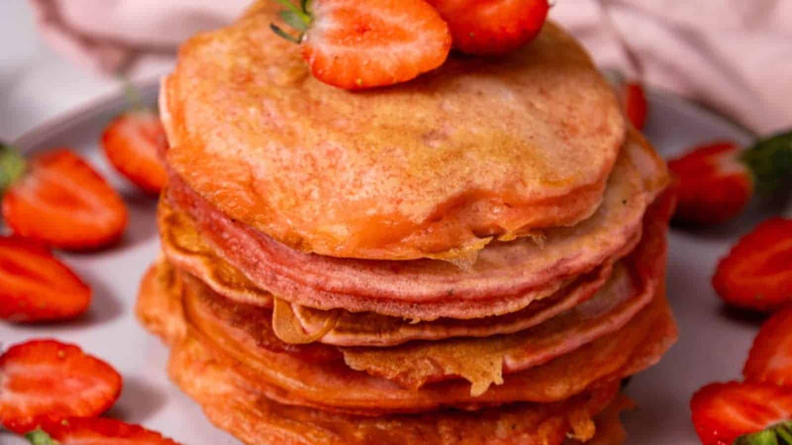 A stack of pancakes topped with sliced strawberries, surrounded by more strawberries on a plate.