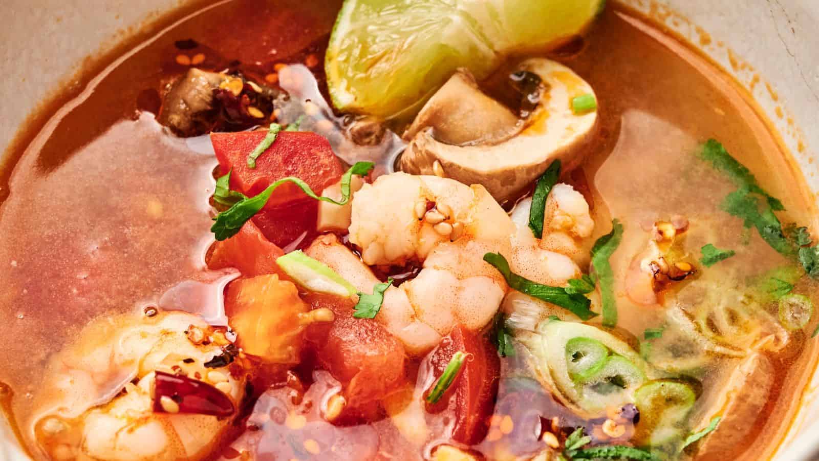 Close-up of a spicy tom yum soup with shrimp, mushrooms, tomato chunks, lime wedge, and chopped herbs in a rich, red broth.