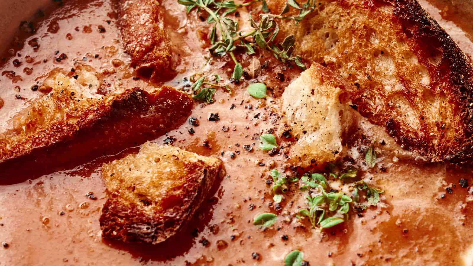 Close-up of a bowl of roasted tomato soup topped with toasted bread chunks, fresh thyme, and a sprinkle of black pepper.