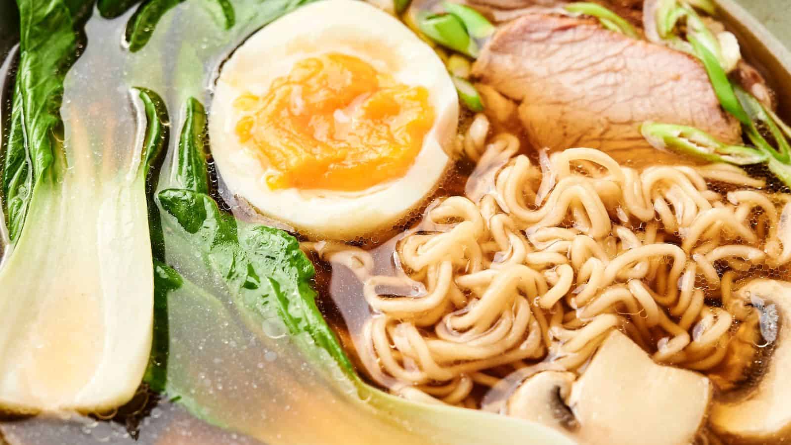 Close-up of a bowl of ramen with noodles, a halved boiled egg, sliced meat, green leafy vegetables, and mushrooms in broth.