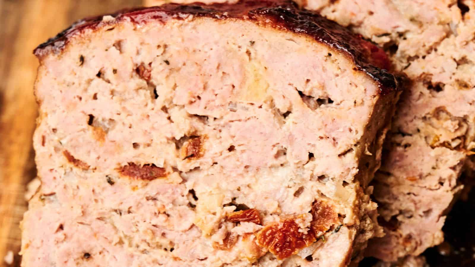 Close-up view of sliced turkey meatloaf with a browned crust on top, revealing a moist and textured interior. The meatloaf appears to contain pieces of onions and possibly sun-dried tomatoes.