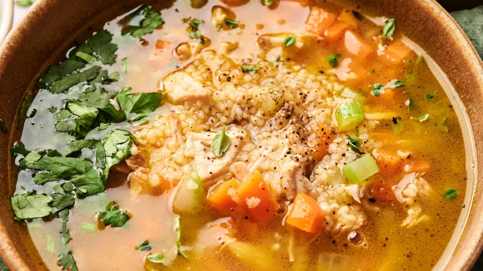 A bowl of chicken soup with rice, carrots, celery, herbs, and pepper sprinkled on top.