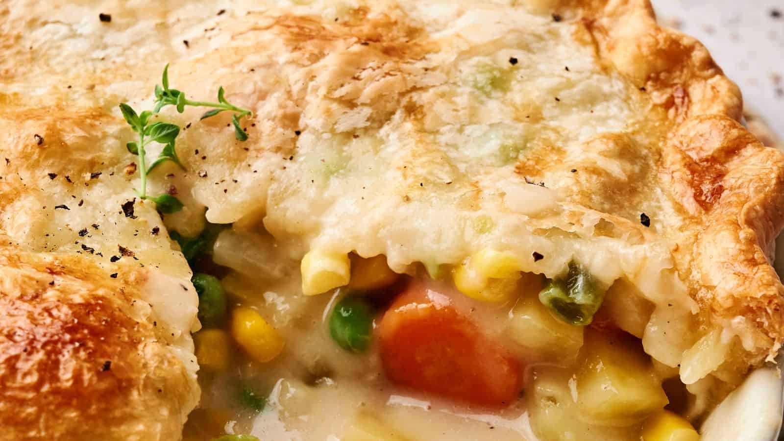 Close-up of a vegan pot pie with a flaky crust, featuring visible corn, peas, carrots, and a creamy filling. A sprig of fresh herb and a sprinkle of black pepper garnish the pie.