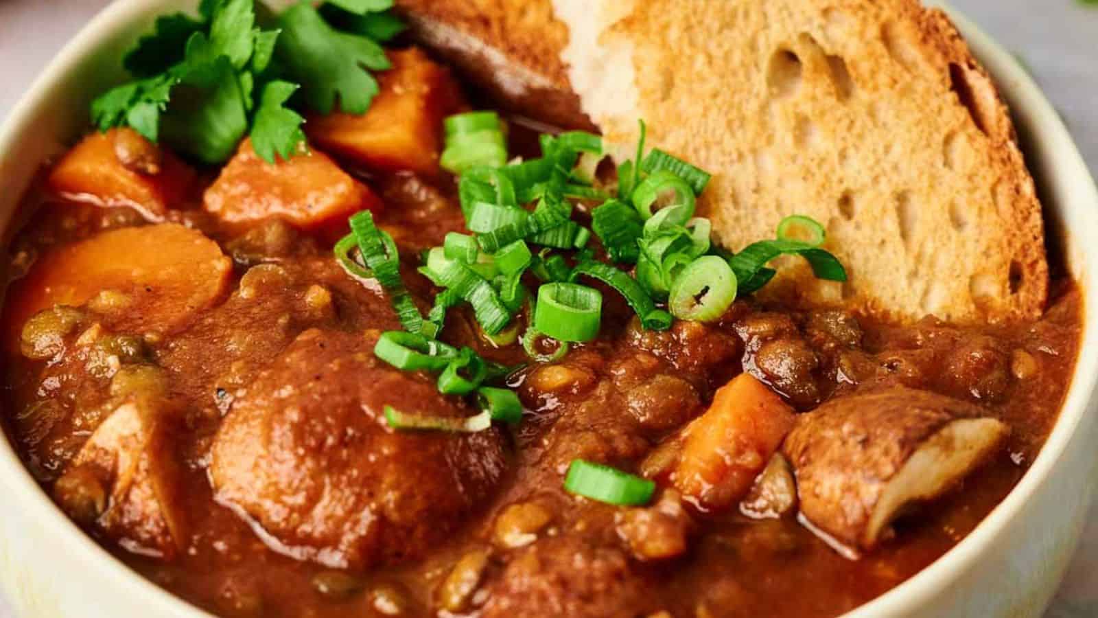 A bowl of lentil stew with potatoes and carrots, garnished with sliced green onions and parsley, accompanied by slices of crusty bread.