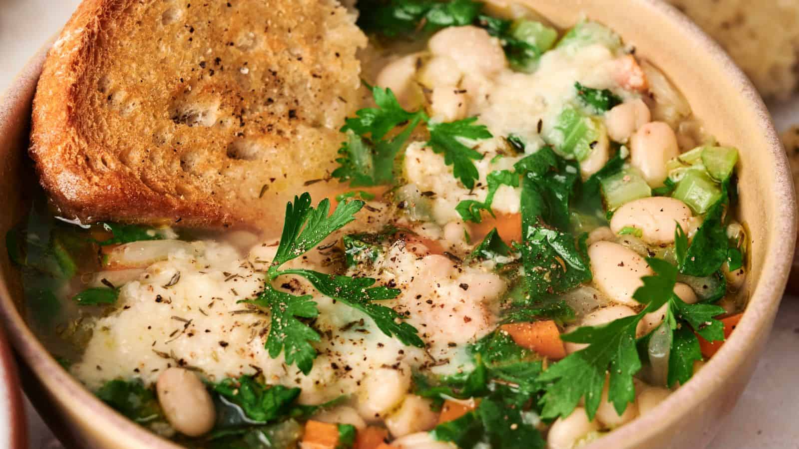 A bowl of hearty white bean soup topped with melted cheese and a slice of toasted bread, garnished with fresh parsley.