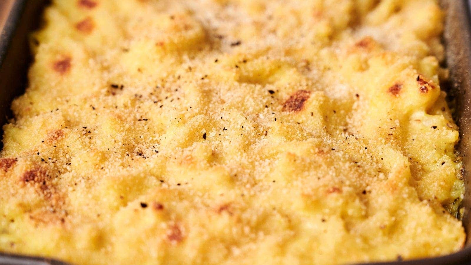 Close-up of a creamy, golden-brown macaroni and cheese dish with a crunchy breadcrumb topping reminiscent of comforting funeral potatoes. A spoon is partially submerged in the cheese sauce.