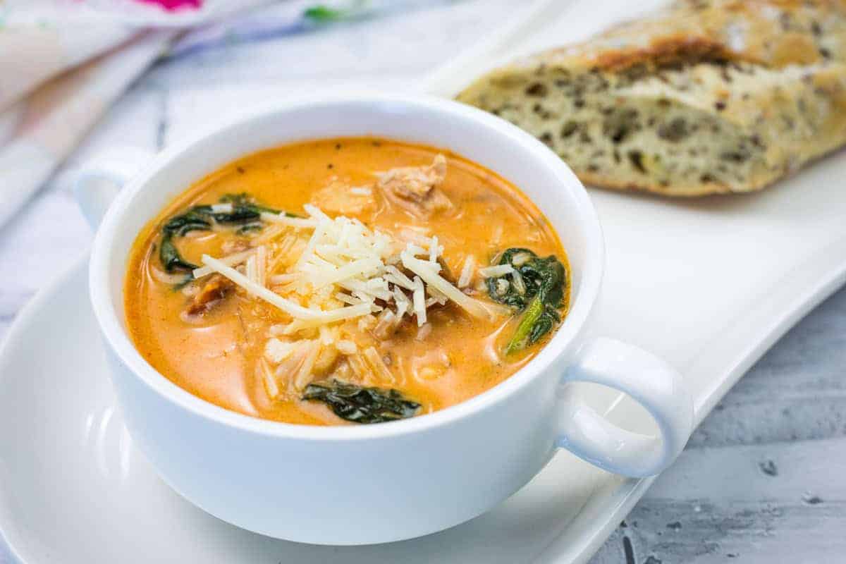 A bowl of Marry Me Chicken Soup accompanied by a piece of bread on a white plate.