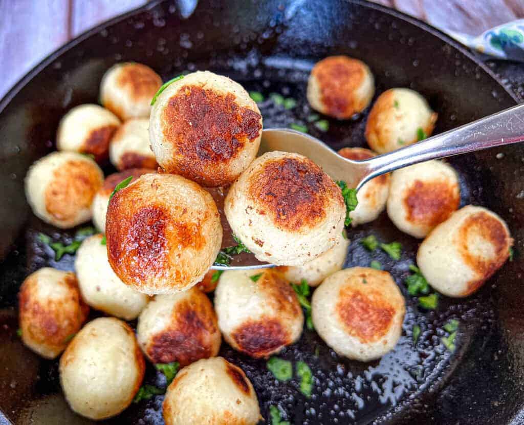 A spoon lifts several Parisian potatoes from a black skillet filled with more Parisian potatoes.