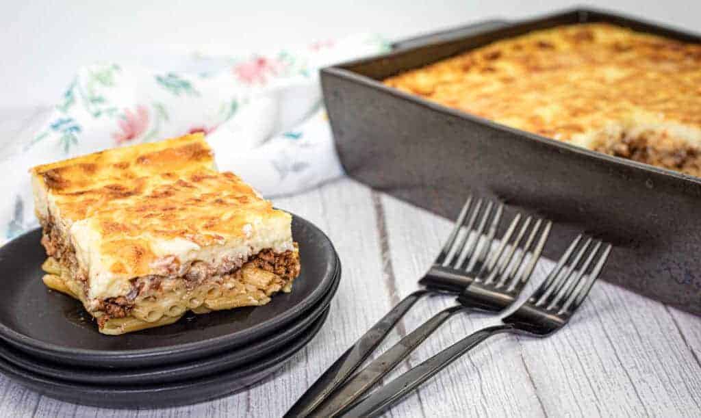 A square piece of pastitsio served on a stack of black plates with three forks and a rectangular baking tray filled with the same pasta on the side.