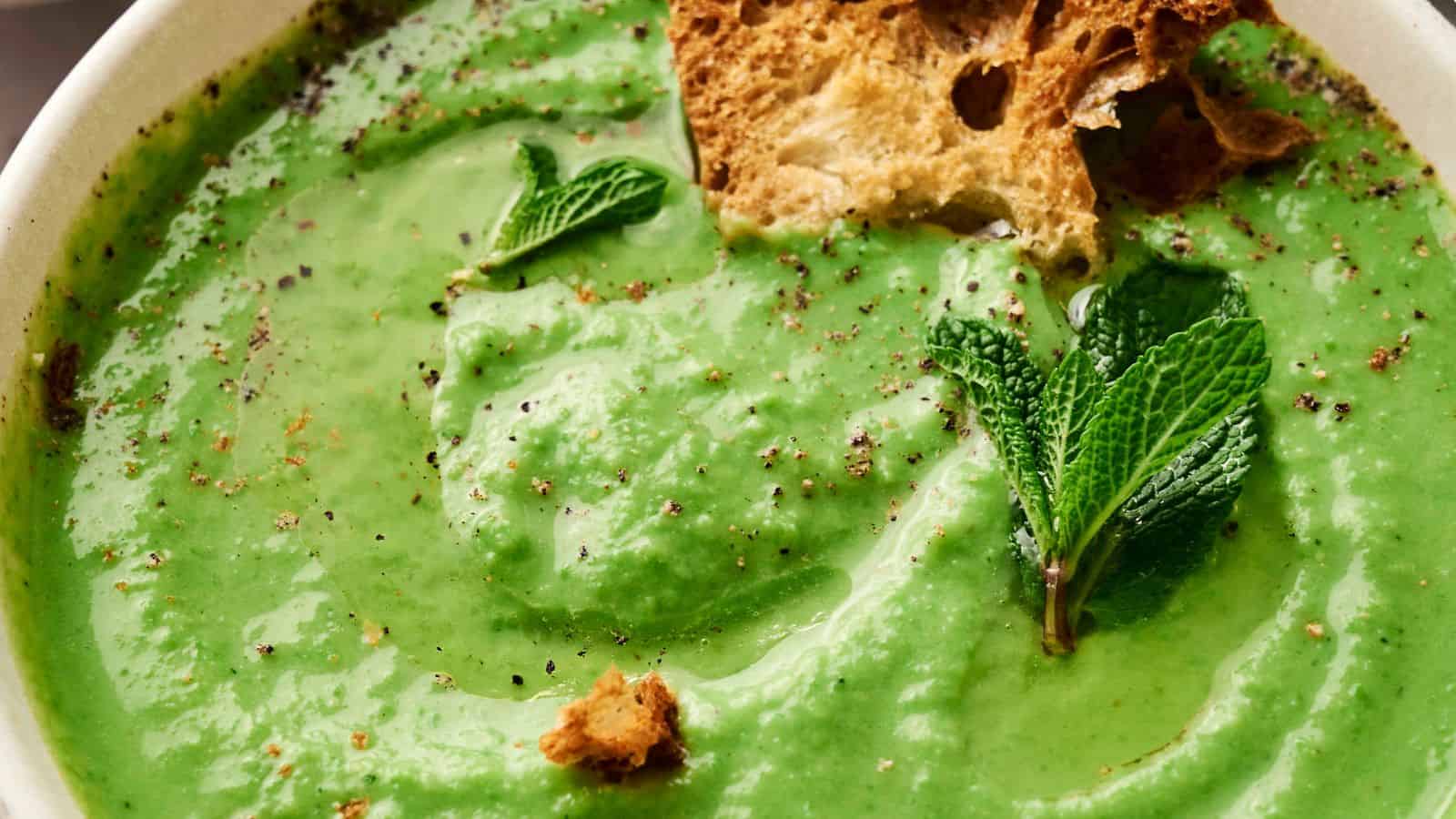 A bowl of creamy green pea soup garnished with mint leaves, black pepper, and pieces of crusty bread.