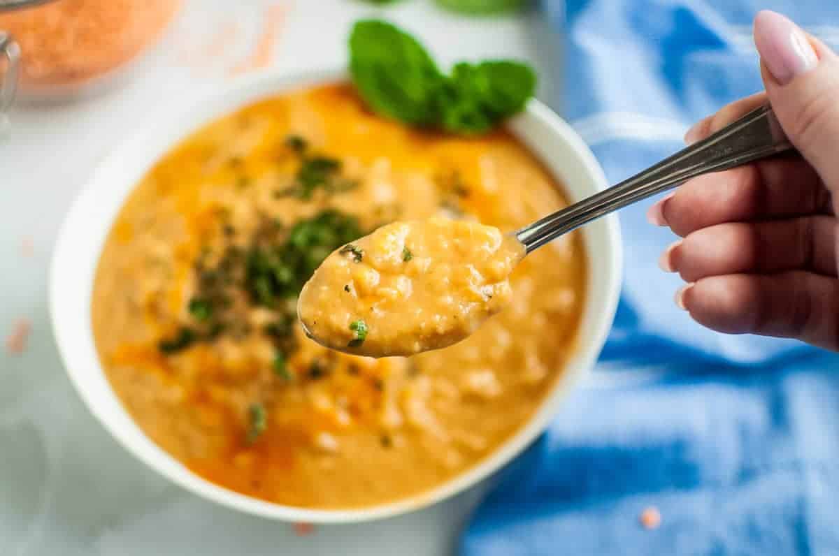 A spoon lifts a spoonful of red lentil soup from a white bowl.