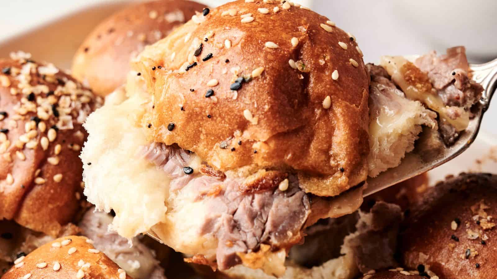 Close-up of a fork lifting a beef sandwich topped with sesame seeds on a soft bun, with more sandwiches in the background.