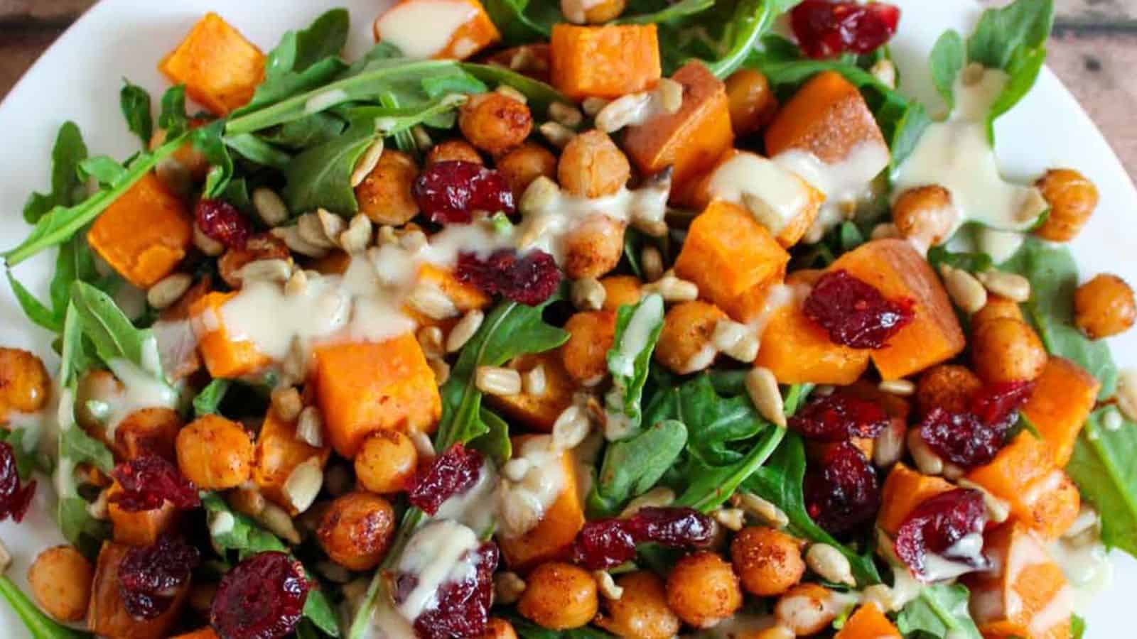 A salad with arugula, roasted sweet potatoes, chickpeas, dried cranberries, sunflower seeds, and a drizzle of creamy dressing on a white plate.