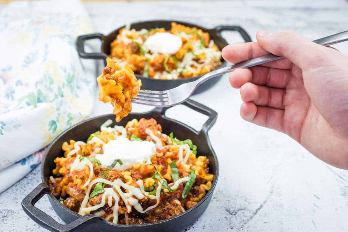 A hand holds a fork above a skillet of lasagna in a black bowl.