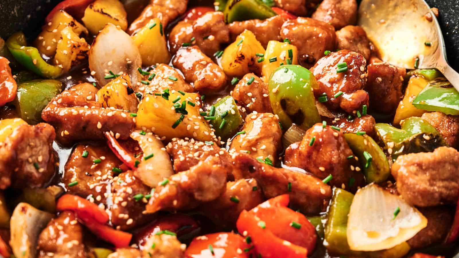 Close-up of sweet and sour pork with chunks of bell peppers, onions, and pineapple, garnished with sesame seeds and chives.