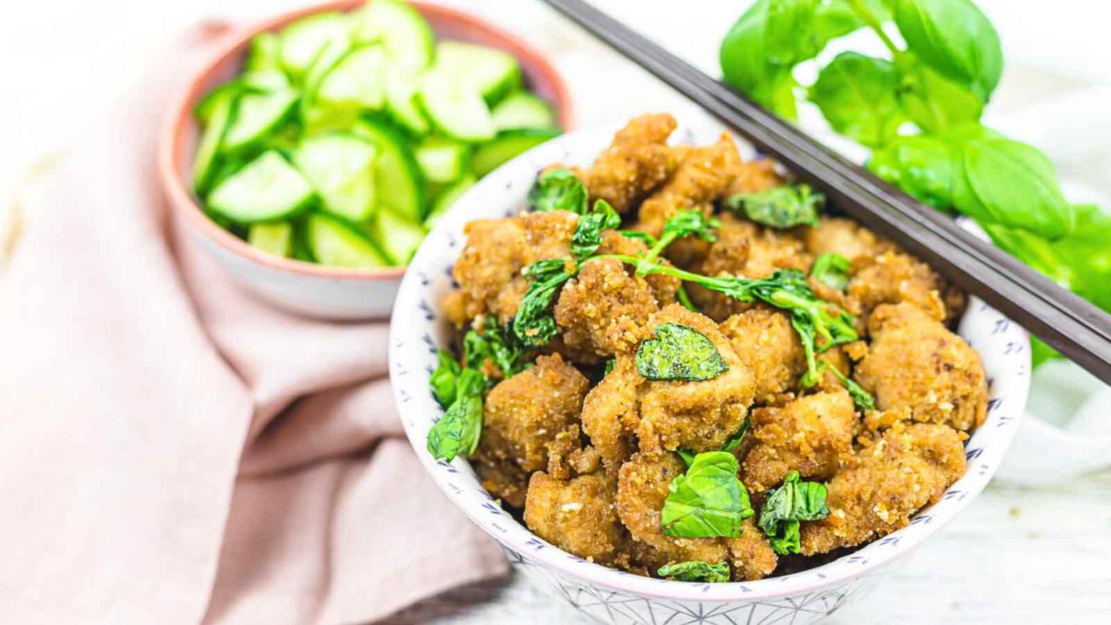 A bowl of crispy fried chicken with fresh basil leaves, accompanied by a side of sliced cucumbers and a basil in the background.