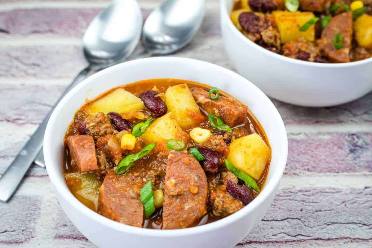 Bowl of Texas cowboy stew accompanied by spoons, on a brick-patterned surface.