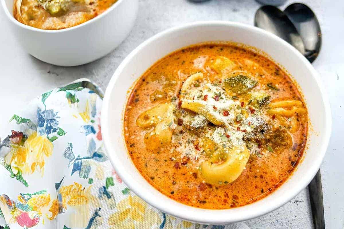 A bowl of creamy tortellini soup is topped with grated cheese and herbs, accompanied by a floral napkin and two spoons on the side.