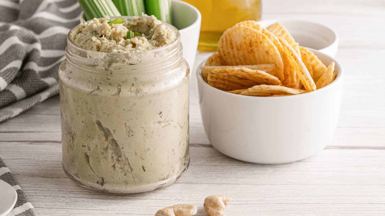 A jar of vegan cashew cheese spread with chives, served with potato chips and celery on a wooden surface.