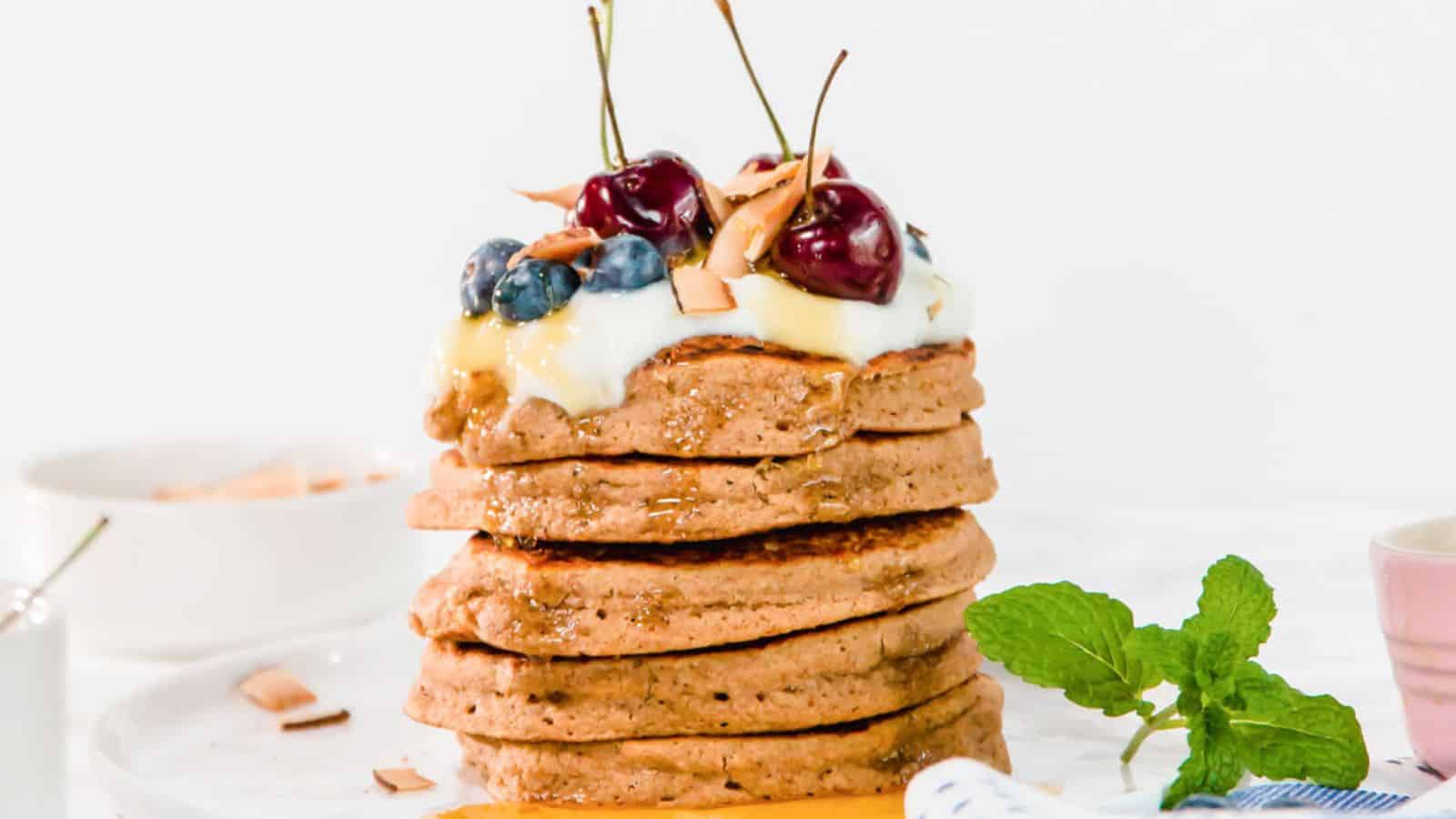 A stack of vegan oatmeal pancakes topped with yogurt, cherries, blueberries, and coconut flakes.