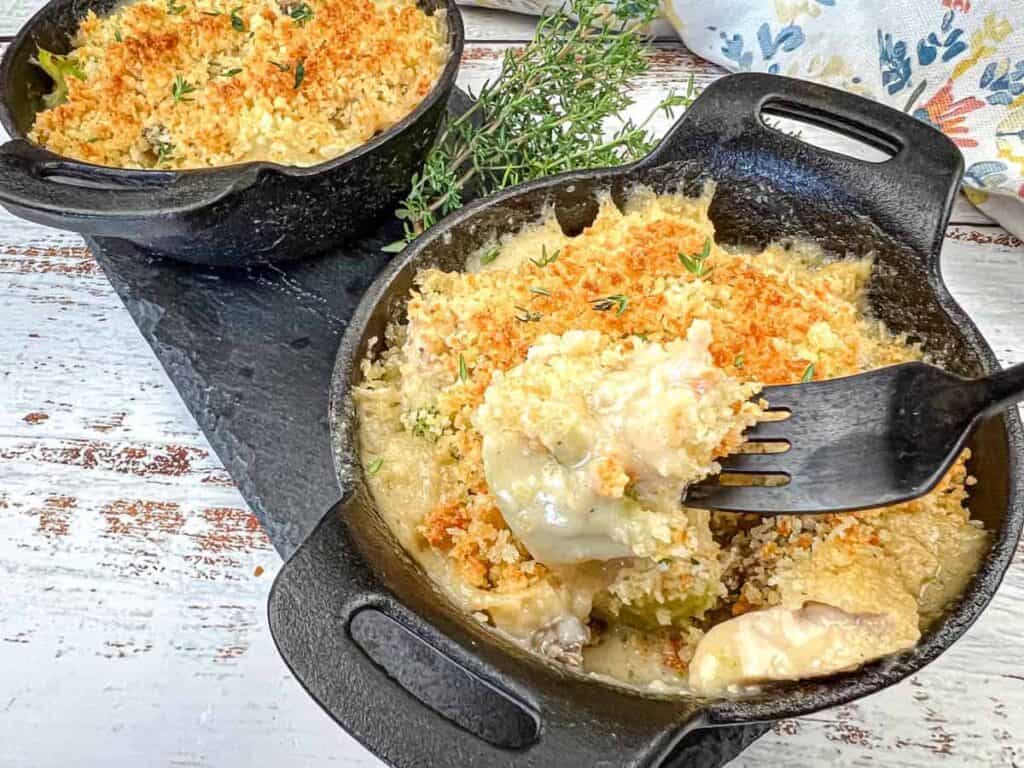 Two cast-iron skillets filled with vintage chicken divan and a fork dishing up one serving.