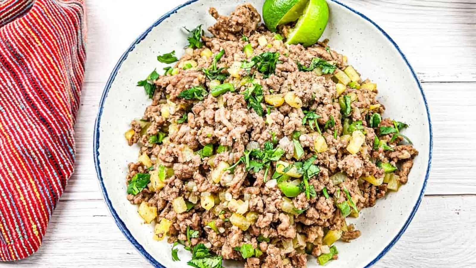 A plate of cooked ground beef mixed with diced vegetables and garnished with lime wedges and chopped herbs. A red striped cloth is placed beside the plate.