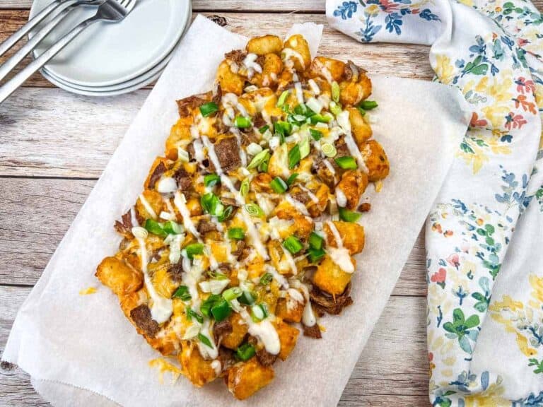 Philly Cheesesteak Loaded Tots on parchment paper, topped with Alfredo cheese, green onions, and a drizzle of white sauce, placed on parchment paper.