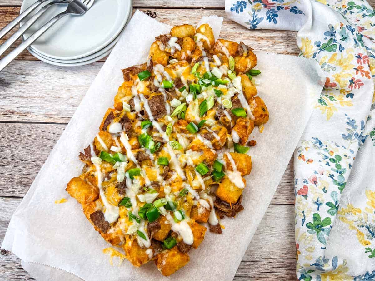 Philly Cheesesteak Loaded Tots on parchment paper, topped with Alfredo cheese, green onions, and a drizzle of white sauce, placed on a dark rectangular slate plate.