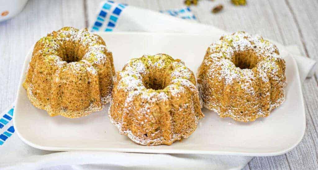 Three pistachio olive oil cakes topped with powdered sugar are arranged on a white rectangular plate.