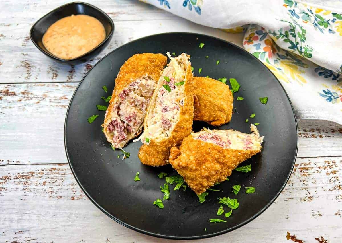 A black plate with Reuben Egg Rolls and dipping sauce.