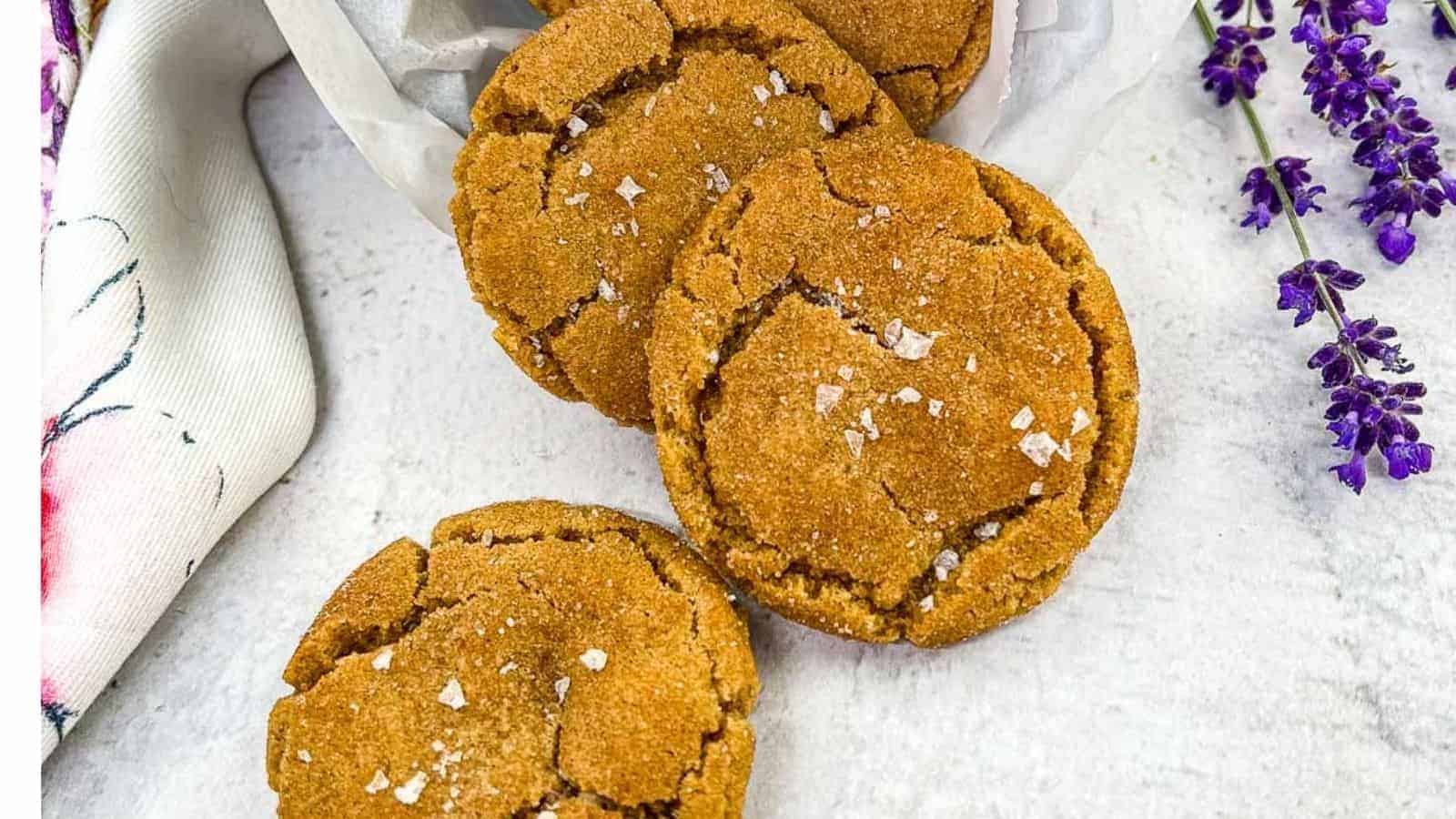 Three brown butter snickerdoodle cookies arranged on a white surface, sprinkled with sea salt flakes, with a white cloth and purple flowers on the side.