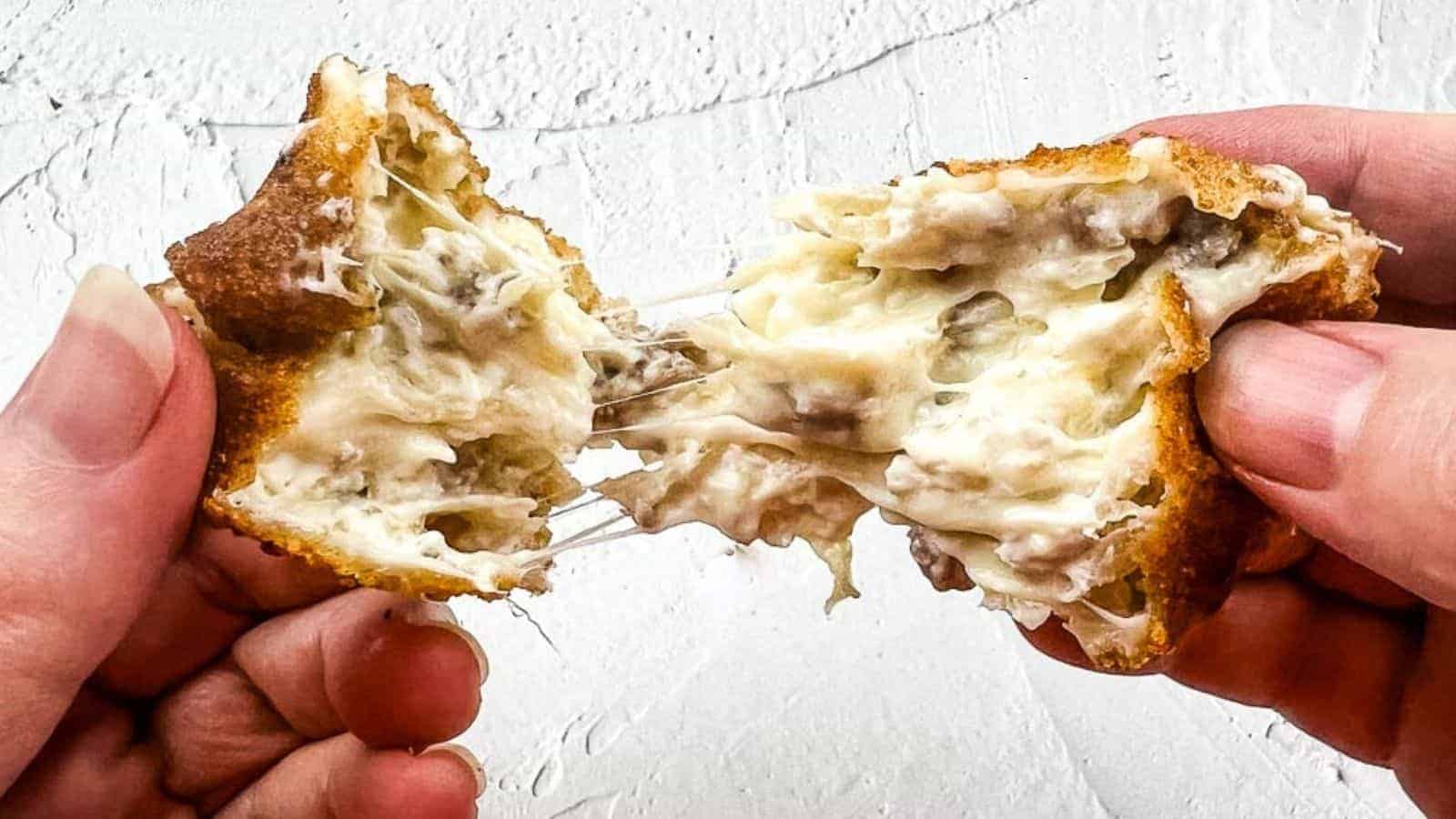 A close-up shot of hands pulling apart a crispy, golden-brown, cheese-filled snack with a gooey, melted cheese center against a textured white background.