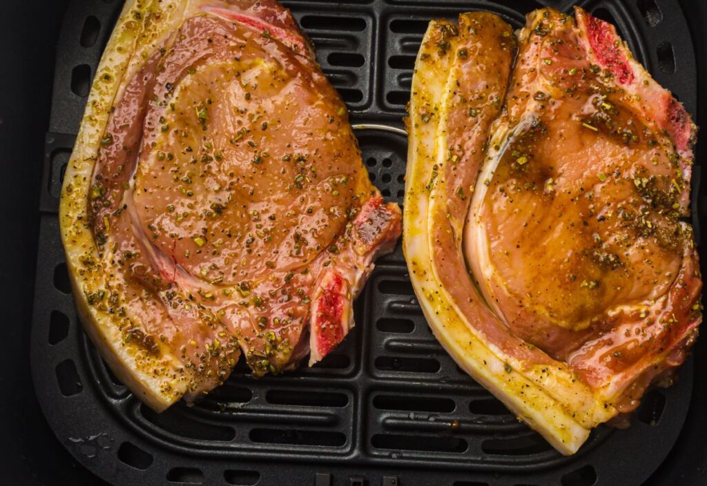 Two seasoned pork chops with visible bone, resting in a black air fryer basket. The chops are coated in a golden-brown marinade speckled with herbs, visible against the pinkish-red meat. The air fryer basket has a grid pattern.