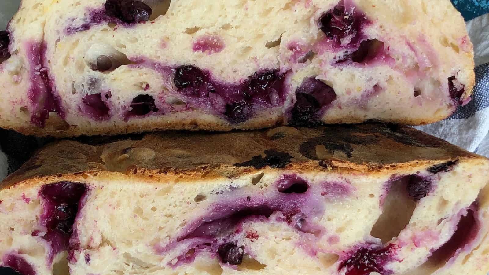 Close-up of two slices of freshly baked bread with visible blueberries and swirls of blueberry juice within the soft crumb. The crust appears lightly browned.