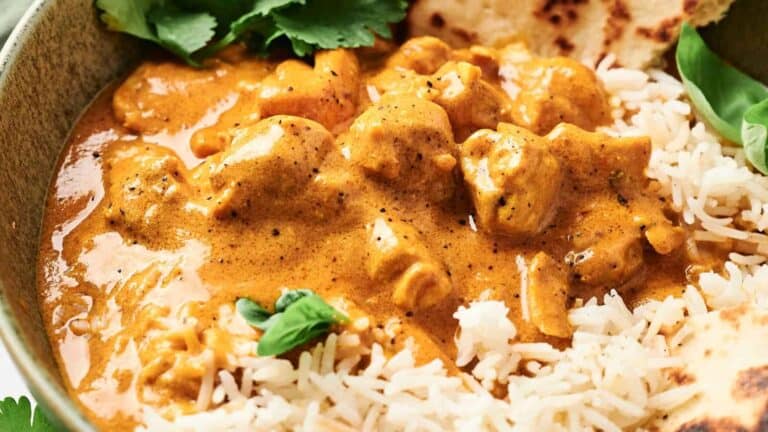 A bowl of creamy curry with chicken pieces served over white rice, garnished with cilantro, and accompanied by flatbread.