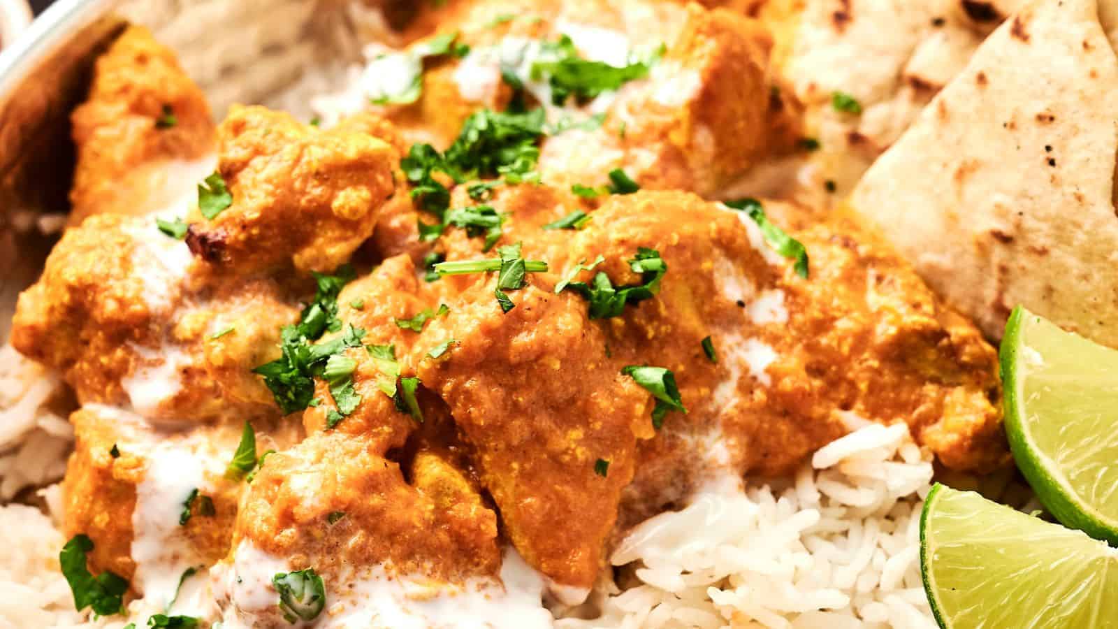 A close-up of chicken tikka masala served on rice, garnished with fresh cilantro, with lime wedges and naan on the side.