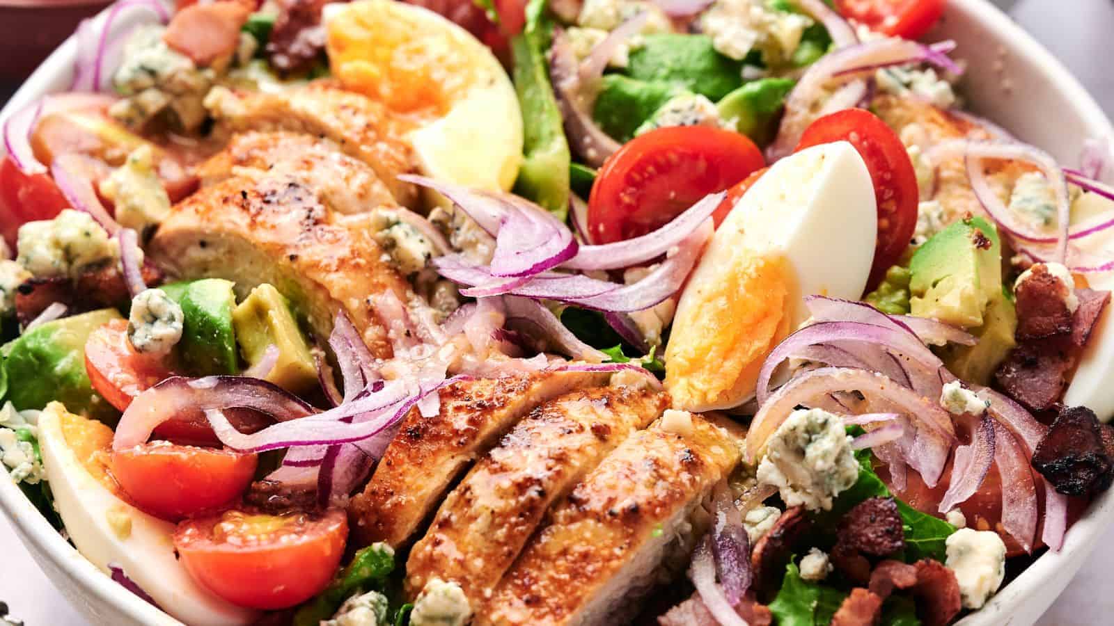 A close-up of a Cobb salad featuring sliced chicken, cherry tomatoes, avocado, boiled eggs, red onion, bacon bits, and blue cheese crumbles on a bed of lettuce.